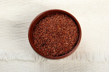 raw red rice in a clay bowl on a light fabric background. uncooked brown wild rice, top view 