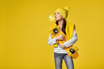 cool hipster little child girl with yellow skateboard blowing a big bubble gum over yellow...