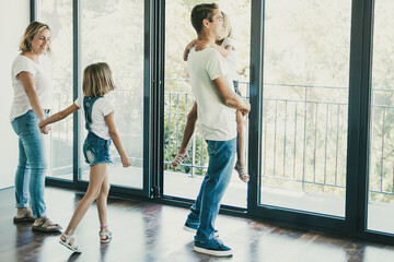 Happy father with daughter standing near open balcony and smiling. Blonde mother holding girl hand and talking with her. Enjoying family watching new house or flat. Relocation and mortgage concept