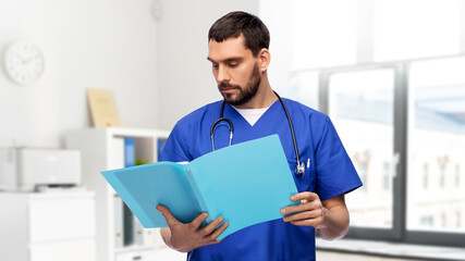 healthcare, profession and medicine concept - doctor or male nurse in blue uniform reading medical report in folder over medical office at hospital on background