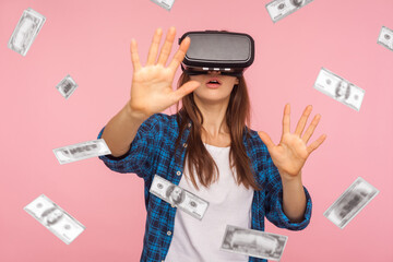 Portrait of shocked woman wearing virtual reality glasses, stretching hands forward to get money, dollar rain falling, illusion of rich millionaire, playing vr game, indoor isolated on pink background