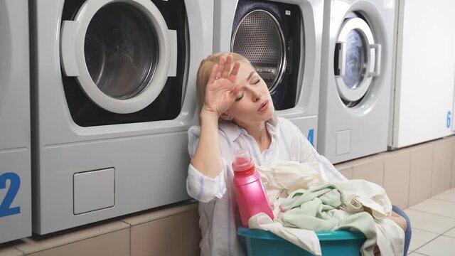 A woman is sitting on the floor of the laundry room next to the washing machines, she is very tired of waiting for her laundry to be washed. A tired woman is waiting for clean clothes in the laundry