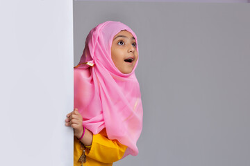 young muslim girl with hijab peeking from behind the wall and looking up in surprise	