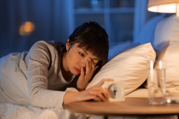 Fototapeta premium people, bedtime and rest concept - sleepy asian woman with clock lying in bed at night