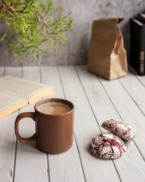 Breakfast with mug of coffee and red velvet cookies. White wooden table, open bookgreen plant and craft package