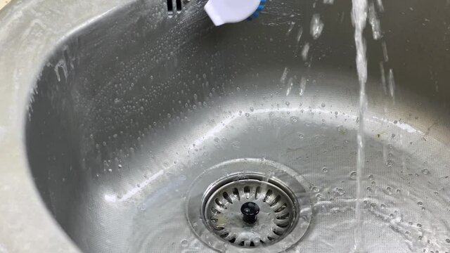 Washing a steel and dirty sink bowl with a brush  under the kitchen krane with a water.