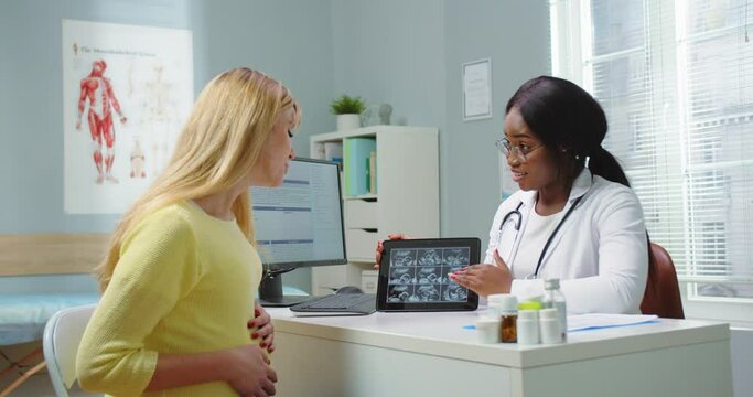 Medium shot of pregnant woman visit african american gynecologist at hospital or medical clinic. Doctor showing tablet with ultrasound pictures to female. Pregnancy and maternity healthcare concept.