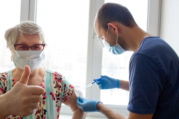 Senior woman in protection mask shows tumbs-up while male doctor giving her an injection Covid-19...