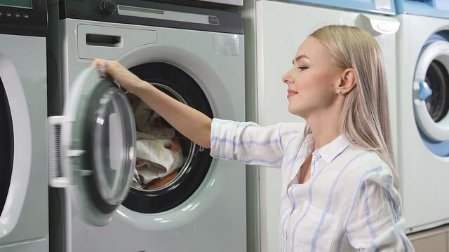 Public self-service laundry. A young pretty woman loads dirty laundry, clothes into the washing machine. slow-motion