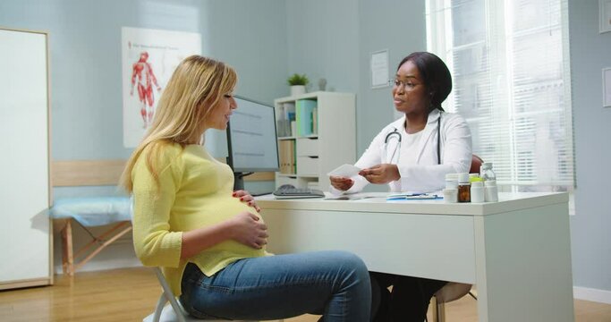 Medium shot of pregnant woman visit african american gynecologist at hospital. Doctor in glasses giving ultrasound picture, baby scan. Pregnancy woman and maternity healthcare concept.