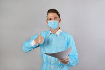 A fair-haired male surgeon in a blue uniform, wearing a medical mask and protective gloves, holds out his documents and shows the OK sign with his hand. The concept of medicine