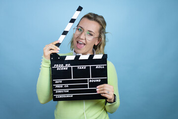Young caucasian woman wearing headphones on neck over blue background holding clapperboard very...