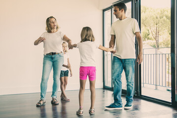 Fototapeta premium Couple of parents and two kids enjoying their new home, standing in empty room and holding hands, dancing. Apartment buying concept