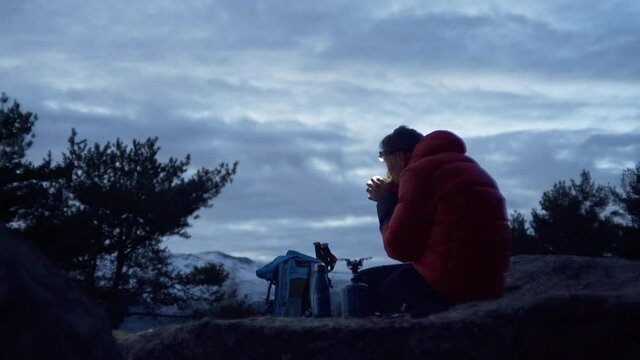 Lonely man on camping trip wears headlamp, look up at sunset sky, sip on coffee or tea prepared on gas camp stove. Adventure in wilderness, urban nomad on travel in nature