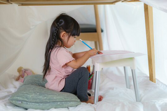 Asian cute little girl drawing in paper while lying in a blanket fort in living room at home for perfect hideout away from their other family members and for them to play imaginatively.