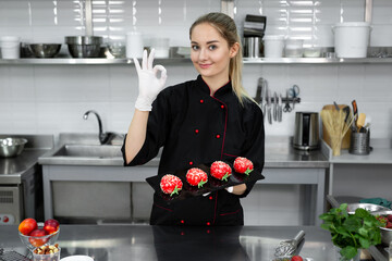 The pastry chef holds a tray with mass cakes, smiles and shows the sign " ok"