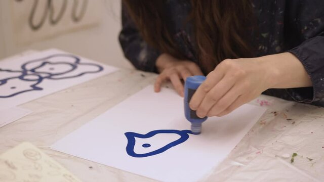 Female artist drawing on a white paper with blue ink. Female painting a bird on a piece of paper, siting to table in art studio, no face