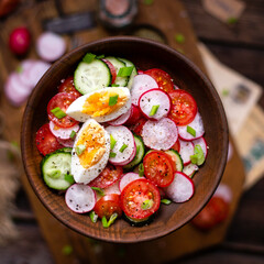 tasty spring salad with round sliced tomatoes, cucumber, radish, boiled eggs, herbs in wooden bowl on rustic table