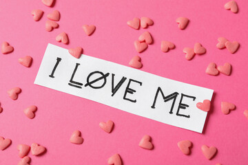 Piece of paper with handwritten phrase I Love Me and heart shaped sprinkles on pink background,...