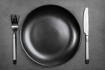 Empty blank black plate with fork and knife on black background of shale serving board. Copy space for prepared dish, recipe or text. Mock up. Top view. Minimal cooking image.