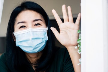 happy Asian woman self-quarantine at home hand waving to someone through window ,social distancing during covid-19 pandemic