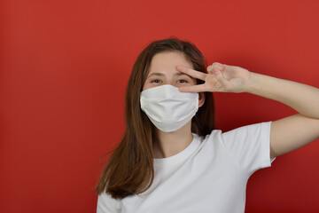 A young girl in a medical mask on a red background. Poster about the coronavirus COVID-19 pandemic. А girl of European appearance in a white t shirt and mask shows a victory sign on a red background