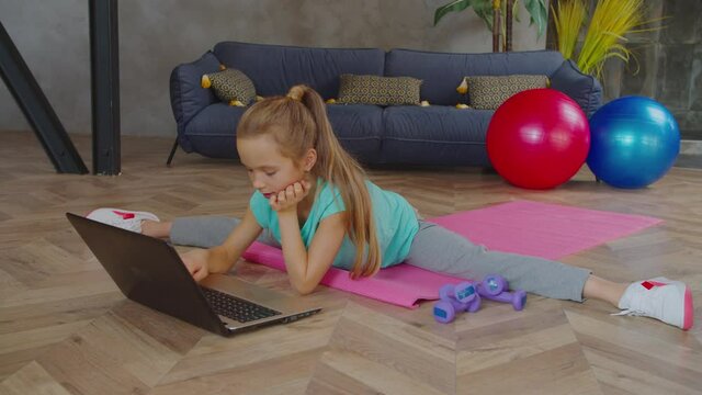 Portrait of flexible adorable fit preadolescent girl sitting in straddle splits on fitness mats, networking online on laptop pc and choosing exercise while practicing sport training in domestic room.