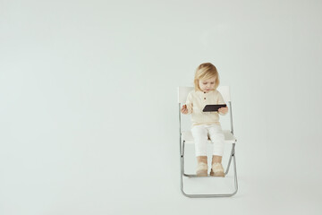 Small child girl sitting on a chair on white isolated background with smartphone in hands eating and watching some cartoons as addiction - 410931629