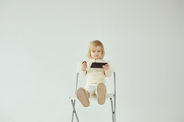 Small child girl sitting on a chair on white isolated background with smartphone in hands eating and watching some cartoons as addiction - 410931836