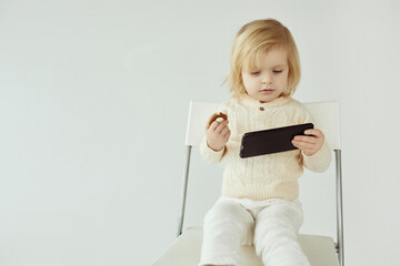 Small child girl sitting on a chair on white isolated background with smartphone in hands eating and watching some cartoons as addiction - 410931870