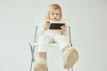 Small child girl sitting on a chair on white isolated background with smartphone in hands eating and watching some cartoons as addiction - 410931889