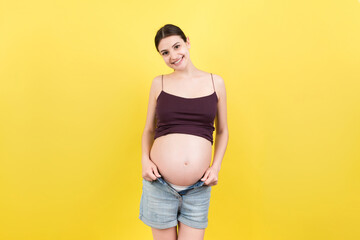Happy pregnant woman in unzipped jeans showing her naked abdomen at colorful background with copy space