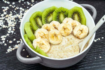 Oatmeal porridge with bananas, kiwi in a bowl with a spoon on a dark background close-up. Healthy breakfast.