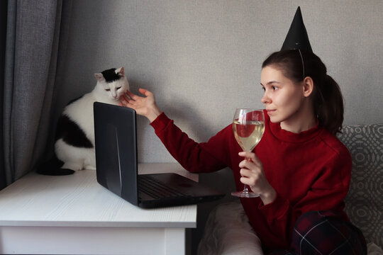 A young woman in a party hat is drinking wine, chatting with someone via video link and stroking a white cat. The person celebrates the holiday while in self-isolation or quarantine due to covid-19