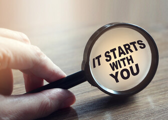 Magnifying glass on wooden table with words quote of It starts with you written on it.