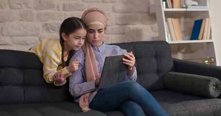 Happy family mother and child daughter in turban are sitting on the sofa, using digital tablet. Cute little girl sitting on sofa using digital tablet with her mother in hijab.