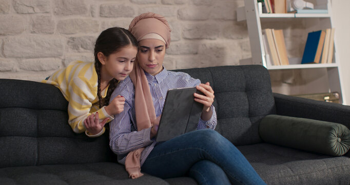 Happy family mother and child daughter in turban are sitting on the sofa, using digital tablet. Cute little girl sitting on sofa using digital tablet with her mother in hijab.