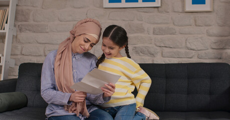 Headscarved mother and daughter are sitting on the sofa and reading letters together. The mother is...
