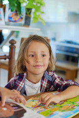 Portrait of joyful little boy which poses sitting at table with books and looks away in living room.