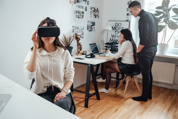 Young businesswoman sitting at table in office room uses virtual reality glasses and enjoys it.