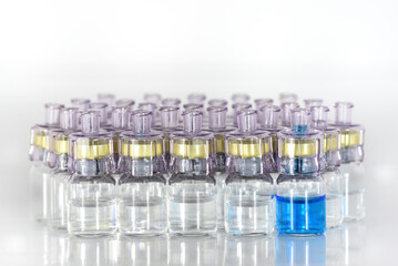 Several ampoules with a transparent liquid, medicine or vaccine stand against a light background. One ampoule contains blue liquid