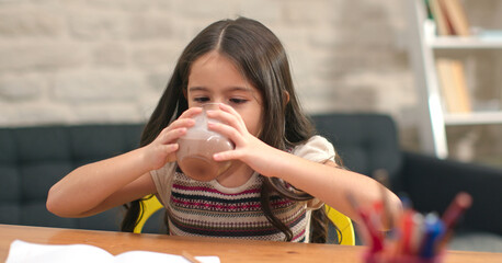 Little girl taking a break from studying, laughing, looking at the camera. Drinking cocoa milk. Distance education of children in quarantine.