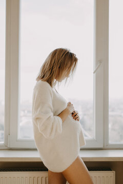 Pregnant woman in a white sweater, standing by the window.