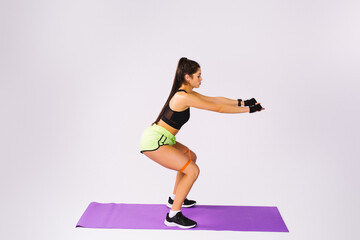 Fototapeta premium Side full-length photo of a young athletic girl who squats with a resistance band. Pumped up buttocks. White background side space for advertising.