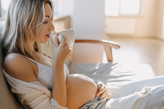 Pregnant woman sitting on a sofa at home, drinking a cup of coffee or tea.