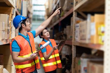 Asian male and female warehouse worker working with clipboard for checking products on shelves shelf pallet in industry storage warehouse. Wearing safety vest and helmet