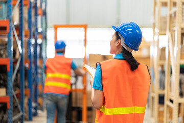 Asian female warehouse worker wearing safety vest and helmet working with clipboard for checking products in parcel on shelves shelf pallet in storage warehouse.  people, warehouse, industry concept