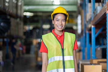 Portrait of smiling black female warehouse worker at work in the industry storage warehouse. African American woman in safety vest and helmet at the shelves shelf warehouse
