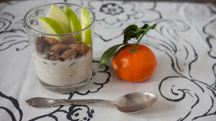 Ovenight oats with nut milk, almonds, granny Smith apple sliced, mandarin. Vintage silver spoon on a white table cloth with ornament.