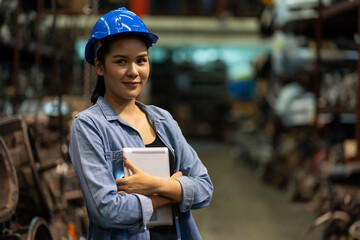 Portrait of Asian female worker in safety uniform and helmet at work in the old automotive spare parts. Women worker holding digital tablet at old garage or automotive spare parts storage warehouse